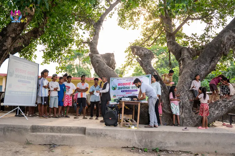 Young people, children and volunteers at an outdoor sanitation awareness programme under a large tree in Rapti Rural Municipality.