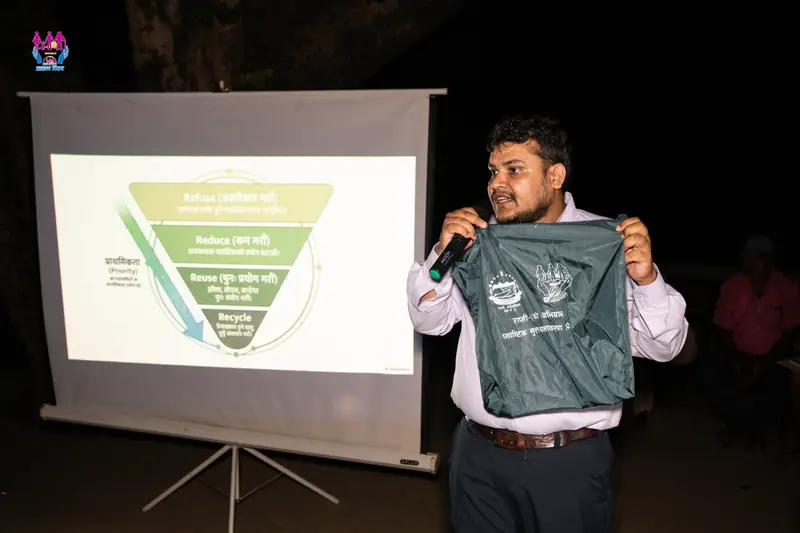 A speaker explains refuse, reduce, reuse and recycle beside a projected slide, holding up a reusable bag, at an evening plastic-free campaign session.